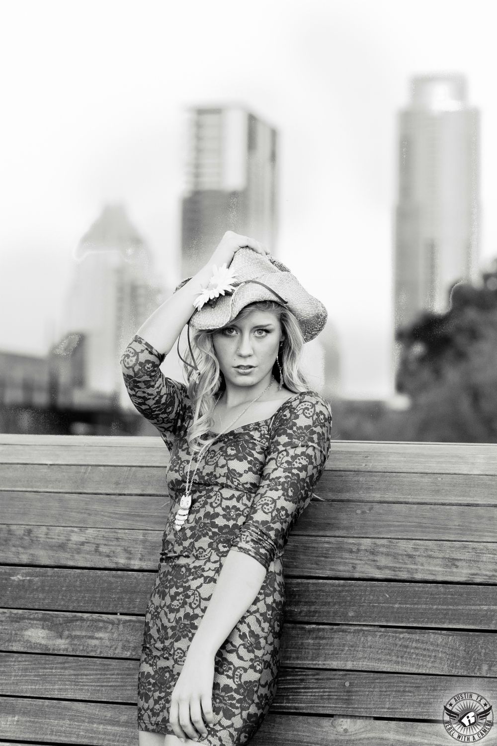 hot blond  fashion model in a lake black and grey skimpy short  hip hugging dress with a cowboy hat in leaning on a wooden rounded railing on the pedestrian bridge over ladybird lake facing West with the skyline of austin out of focus in the background in black and white